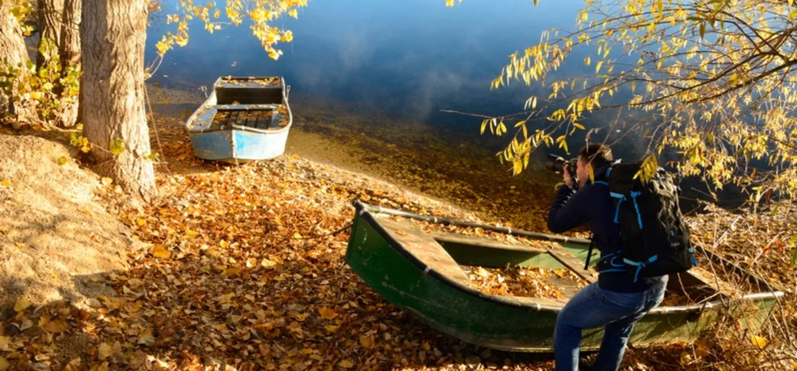 A person with a camera is taking pictures by the lake shore, where there are two old boats—one green and one blue—among autumn leaves and trees by the calm water.