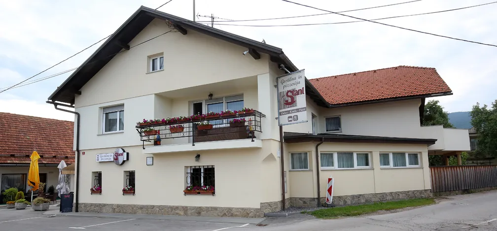 Zunaji izgled stavbe, kjer je gostilna in pizzeria Pri Stani. Enonadstropna hiša bledorumenkaste barve, na balkonu rože.