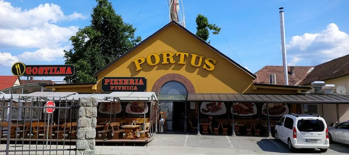 Zunanji izgled gostilne Portus z napisi in rumeno fasado ter ograjo in parkiriščem.