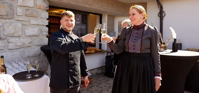 Moški v kuharski uniformi in ženska v tradicionalno navdihnjeni obleki nazdravljata s kozarci penine pred Notranjsko kuhno, ob kamniti steni in pripravljeni pogostitveni mizi.