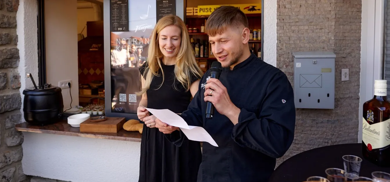 Kuhar govori v mikrofon in bere z lista papirja, ob njem pa stoji nasmejana ženska; za njima je okno Notranjske kuhne z razstavljenimi izdelki.