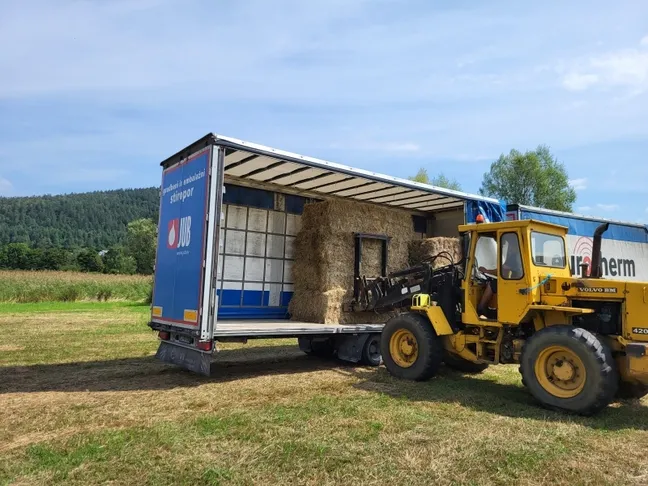 Tovornjak z odprtim priklopnikom, napolnjenim s pravokotnimi balami sena, je parkiran na travnatem polju. Poleg tovornjaka stoji rumeni nakladalnik, ki dviga in naklada bale na priklopnik. V ozadju so drevesa, travniki in gozdnata hribovita pokrajina pod modrim nebom.