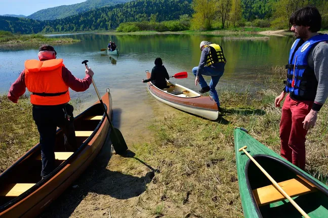 Skupina ljudi ob jezeru pripravlja kanuje za spust; nekateri vstopajo v čolne, drugi stojijo ob obali z rešilnimi jopiči, medtem ko je en kanu že na vodi. V ozadju so gozdnata pobočja in hribi.