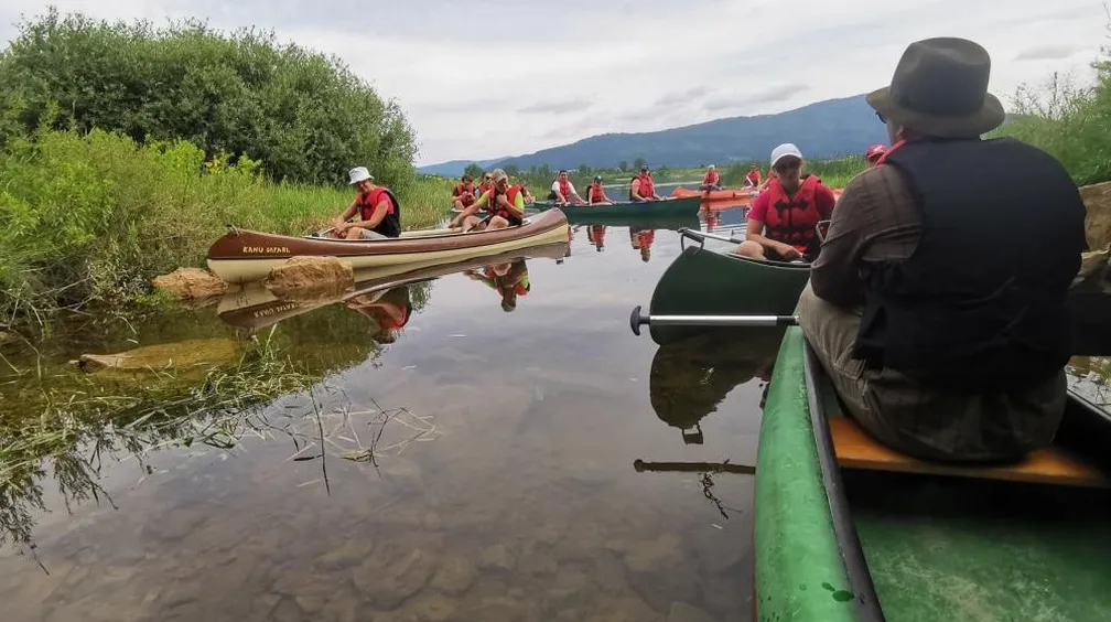 Skupina ljudi v kanujih, ki plujejo pred jamo Skadulco na Cerkniškem jezeru, posluša zgodbo, ki jo pripoveduje vodnik s klobukom.