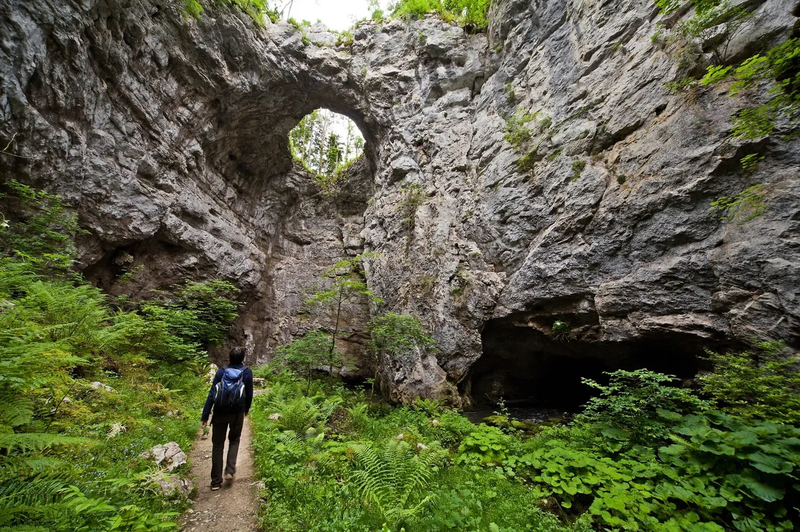 Oseba z nahrbtnikom hodi po ozki poti, obdani z bujnim zelenjem. Pot vodi proti veliki skalni tvorbi z lokasto odprtino na vrhu - Mali naravni most, skozi katero prodira svetloba. Na desni strani slike je viden še en temen vhod v jamo.