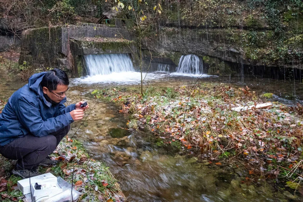 Kitajski raziskovalec, oblečen v moder anorak ter črne hlače, čepi in nekaj gleda. V ozadju vidimo izvir Martinjščice. Pada dež
