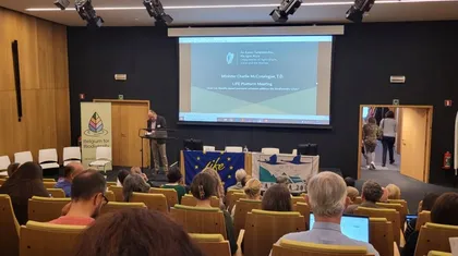 Predavalnica polna ljudi. V centru fotografije je platno, pred njim miza in govorniški pult. Na mizi visijo zastave LIFE in Natura 2000. Za govorniškim pultom je predavatelj.