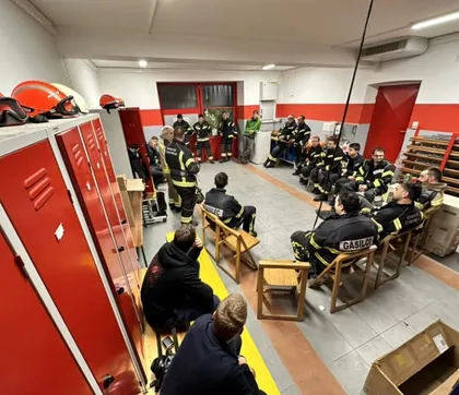 Gasilci oblečeni v svojo uniformo, sedijo na stolih v krogu v Gasilnem domu in se pogovarjajo. Na levi strani so rdeče omarice, na njih oranžne in bele čelade.