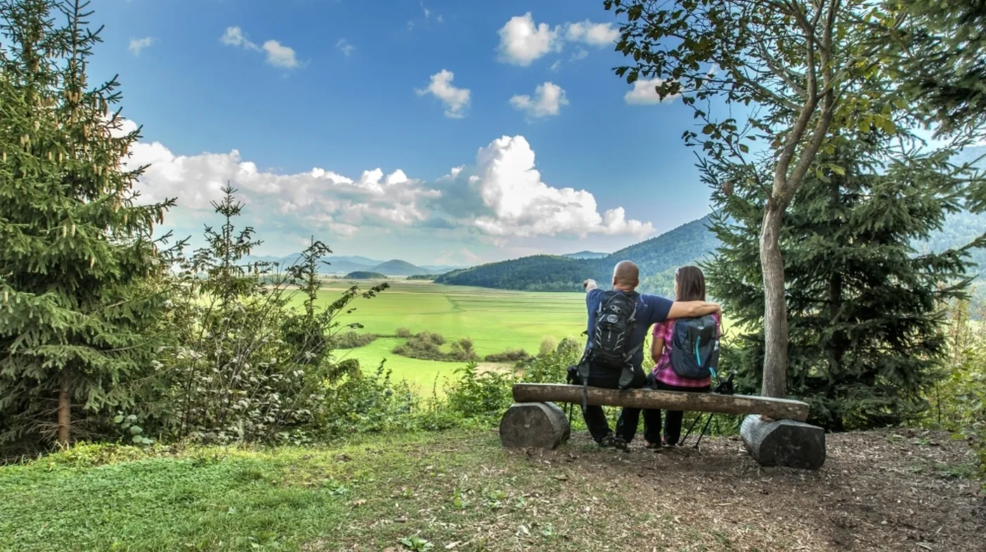 Dve osebi sedita na leseni klopi iz hlodov in opazujeta slikovito pokrajino z zelenimi polji, griči in gorami v daljavi pod delno oblačnim nebom. Klop je postavljena med drevesi, z iglavci na levi in desni strani slike.