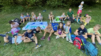 Skupina otrok sedi na travi v naravnem okolju v gozdu. Nekateri sedijo ob zastavi, drugi neposredno na travi. Okoli njih so raztreseni nahrbtniki in drugi predmeti. V ozadju so drevesa in grmovje.