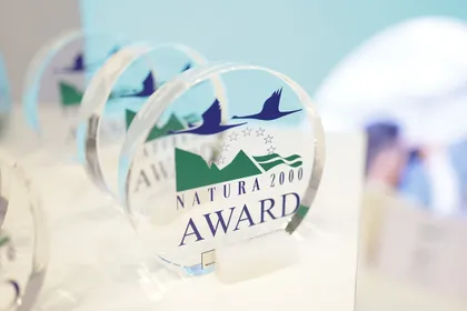 A set of transparent round awards with the inscription 'NATURA 2000 AWARD', decorated with a motif of two flying birds, green mountains, and green waves.