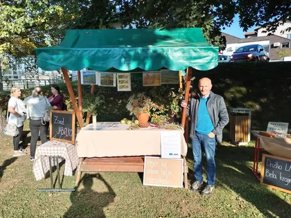 Outdoor market stall with a green canopy, a beige-covered table displaying various items including a vase with flowers. A person stands next to the stall, with signs and boards visible around it.