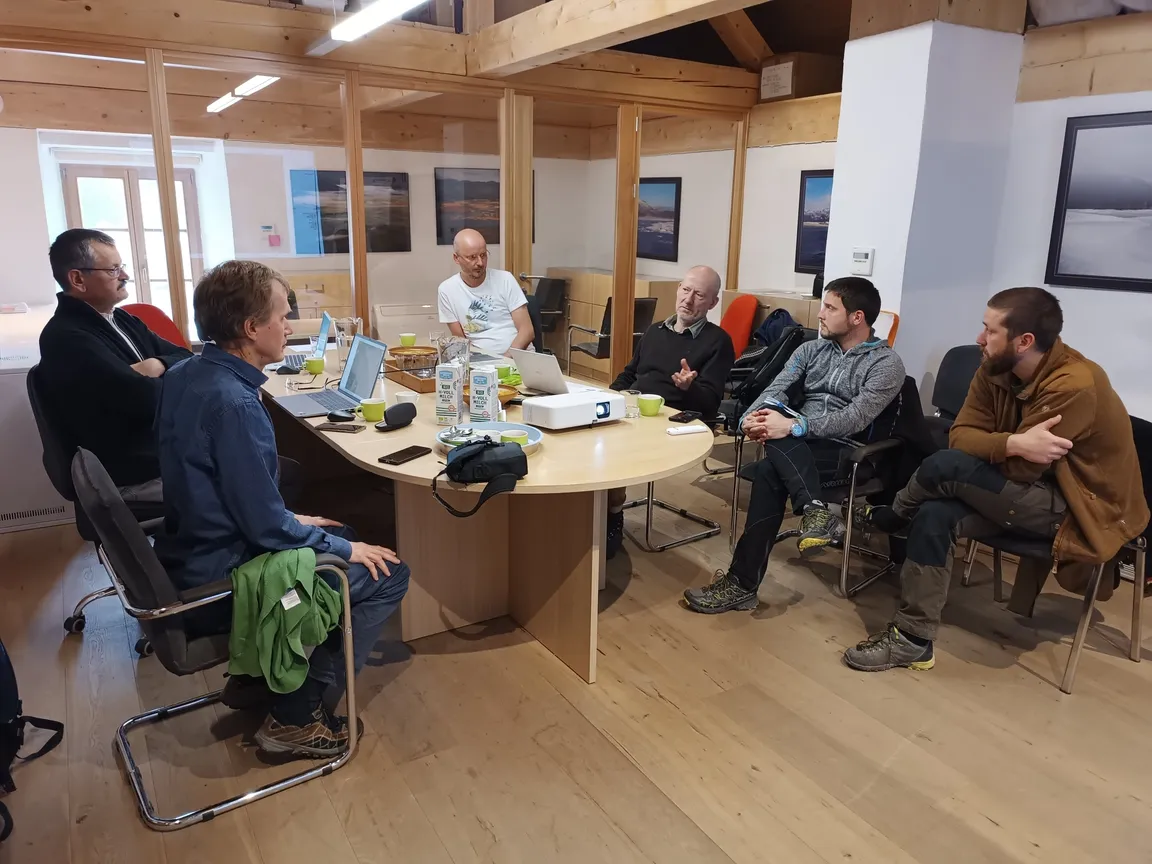 Skupina sedmih ljudi sedi okoli velike ovalne mize v prostoru z lesenimi tlemi in stenami. Na mizi so prenosniki, steklenice z vodo in papirji. Na stenah visijo uokvirjene slike, prostor osvetljuje naravna svetloba skozi okna ter stenske luči.