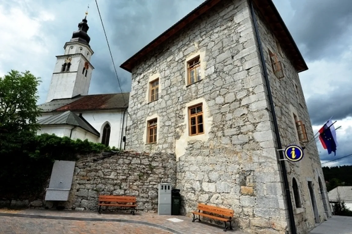 Kamnita stavba z več okni in leseno streho, v ozadju cerkev z visokim zvonikom in uro. Nebo je oblačno, ob kamnitem zidu sta dve klopi. Na desni strani slike sta slovenska zastava in evropska zastava.