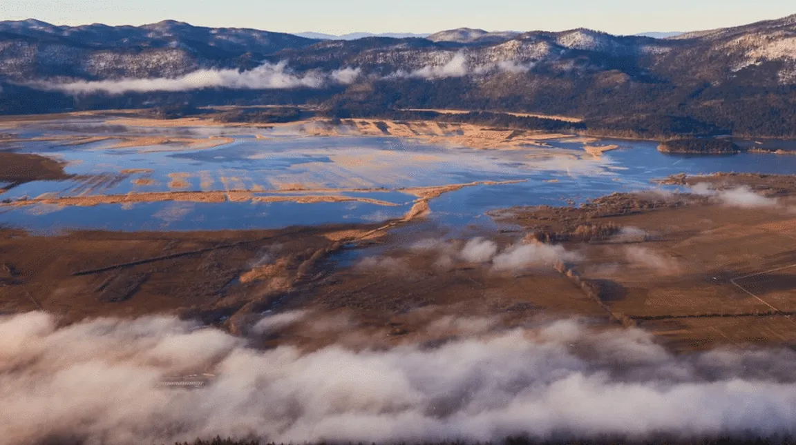 Zračni posnetek pokrajine z velikim vodnim telesom, delno prekritim z ledom, obdan z gorami. V vodi so vidni otočki ali kopenski predeli. Okolica vključuje polja in griče, nekatere površine so prekrite z meglo ali oblaki. Gorski vrhovi v ozadju so zasneženi.