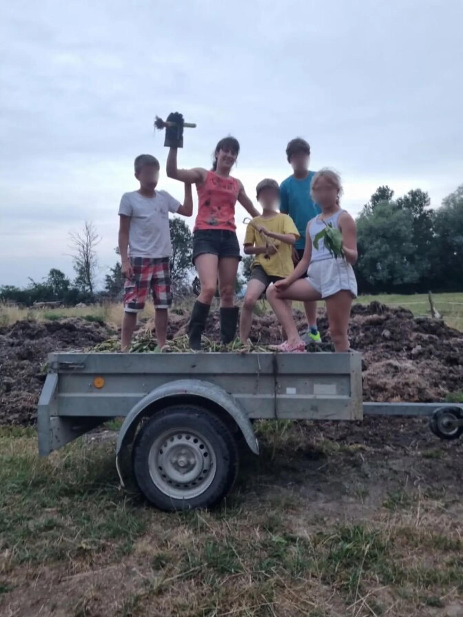 Pet oseb stoji na prikolici, polni zemlje ali komposta; v ozadju polje in drevesa. Ena oseba drži predmet nad glavo, druga rastlino.