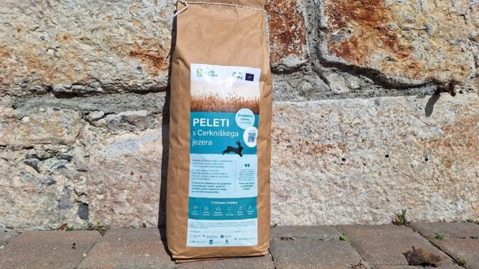 paper bag of pellets labeled 'PELETI s Cerkniškega jezera', placed on a paved surface in front of a stone wall, with a label featuring logos, certifications, and graphics highlighting the product’s natural and eco-friendly composition.