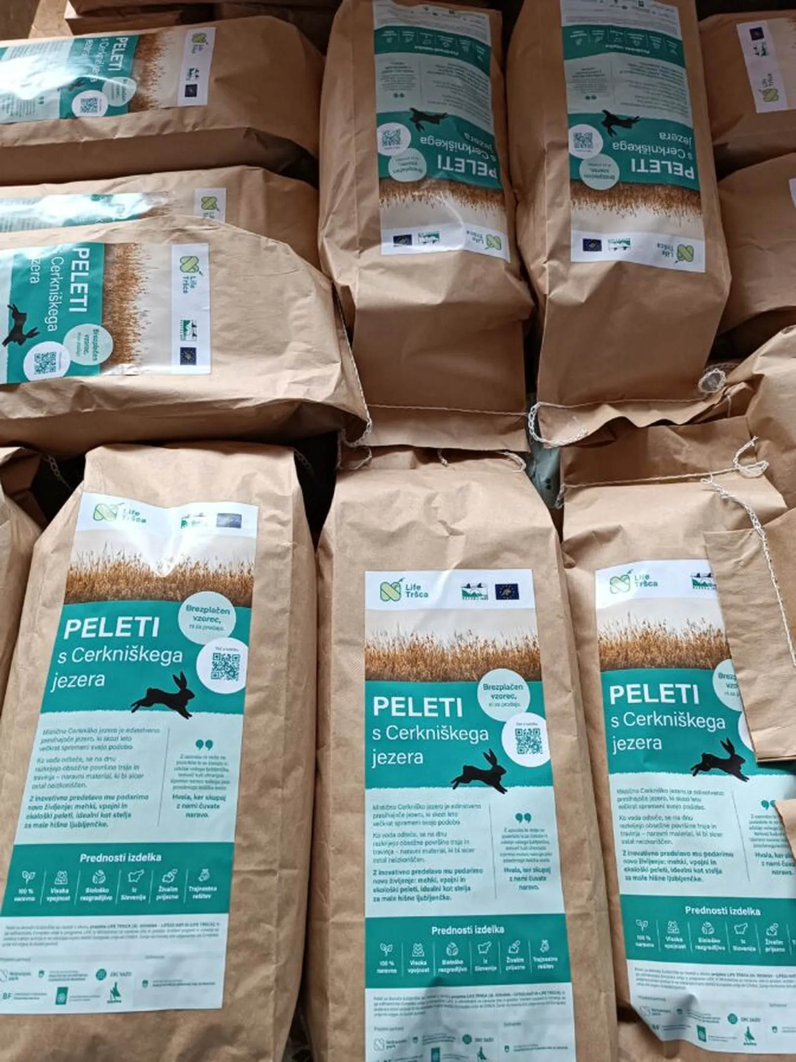 A pile of paper bags, all labeled sticker with 'Peleti s Cerkniškega jezera', featuring logos, a QR code, and icons indicating the product's benefits.