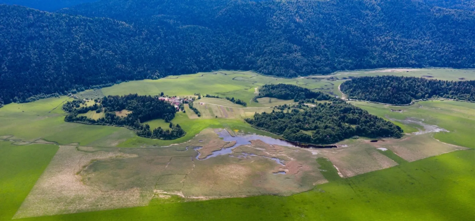 Zračni pogled na zeleno pokrajino z obsežnimi travniki, manjšimi gozdnimi otoki in naseljem na levi strani. V ospredju so mokrišča z vodnimi površinami, v ozadju pa gosta gozdnata pobočja hribov ali gora.