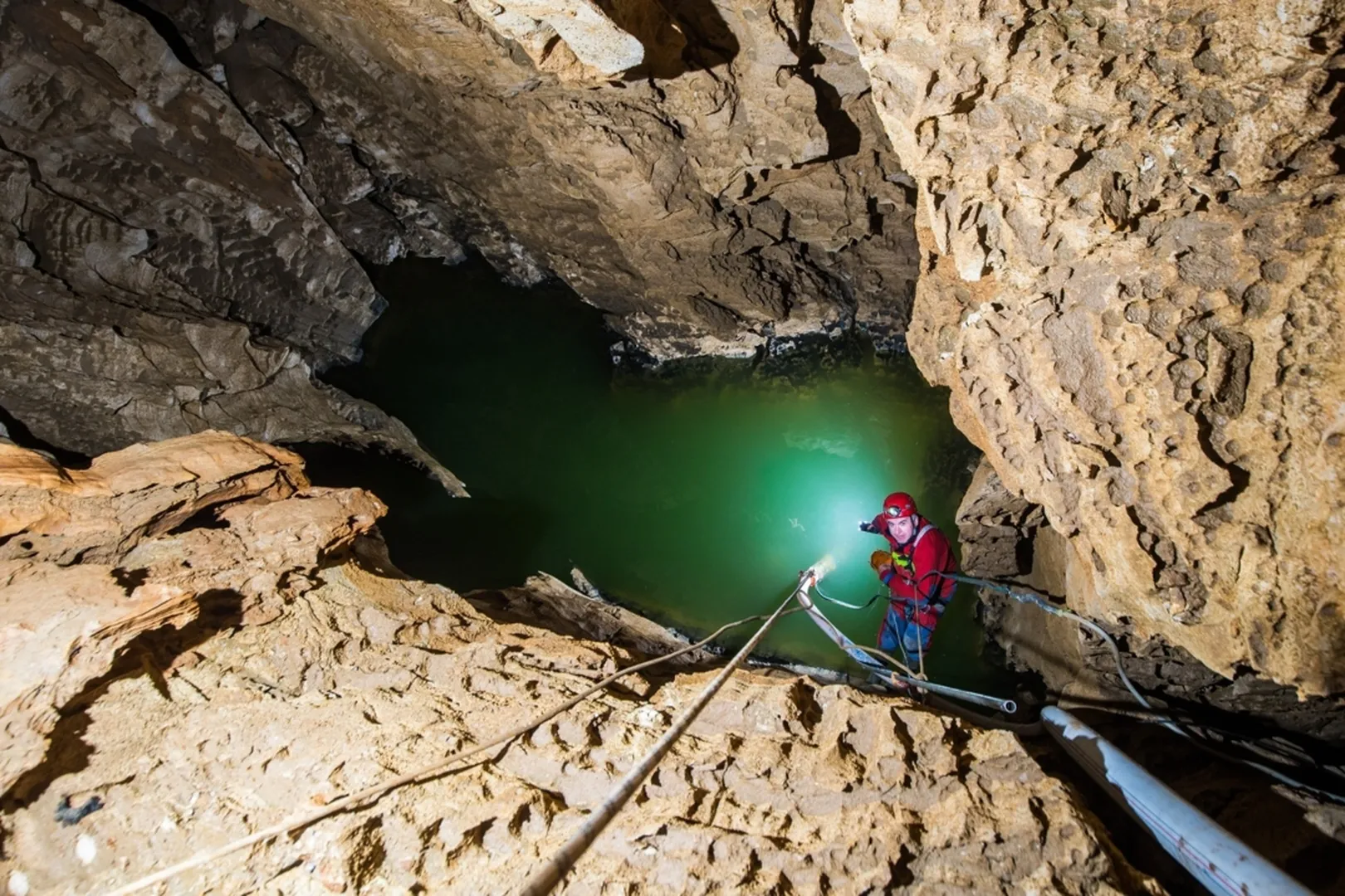 Jamar raziskuje globoko kraško jamo, stoji ob robu strme skalnate stene. Z vrvno opremo in močno svetilko osvetljuje skrivnostno zelenkasto podzemno jezero, ki se skriva v temni globini jame.