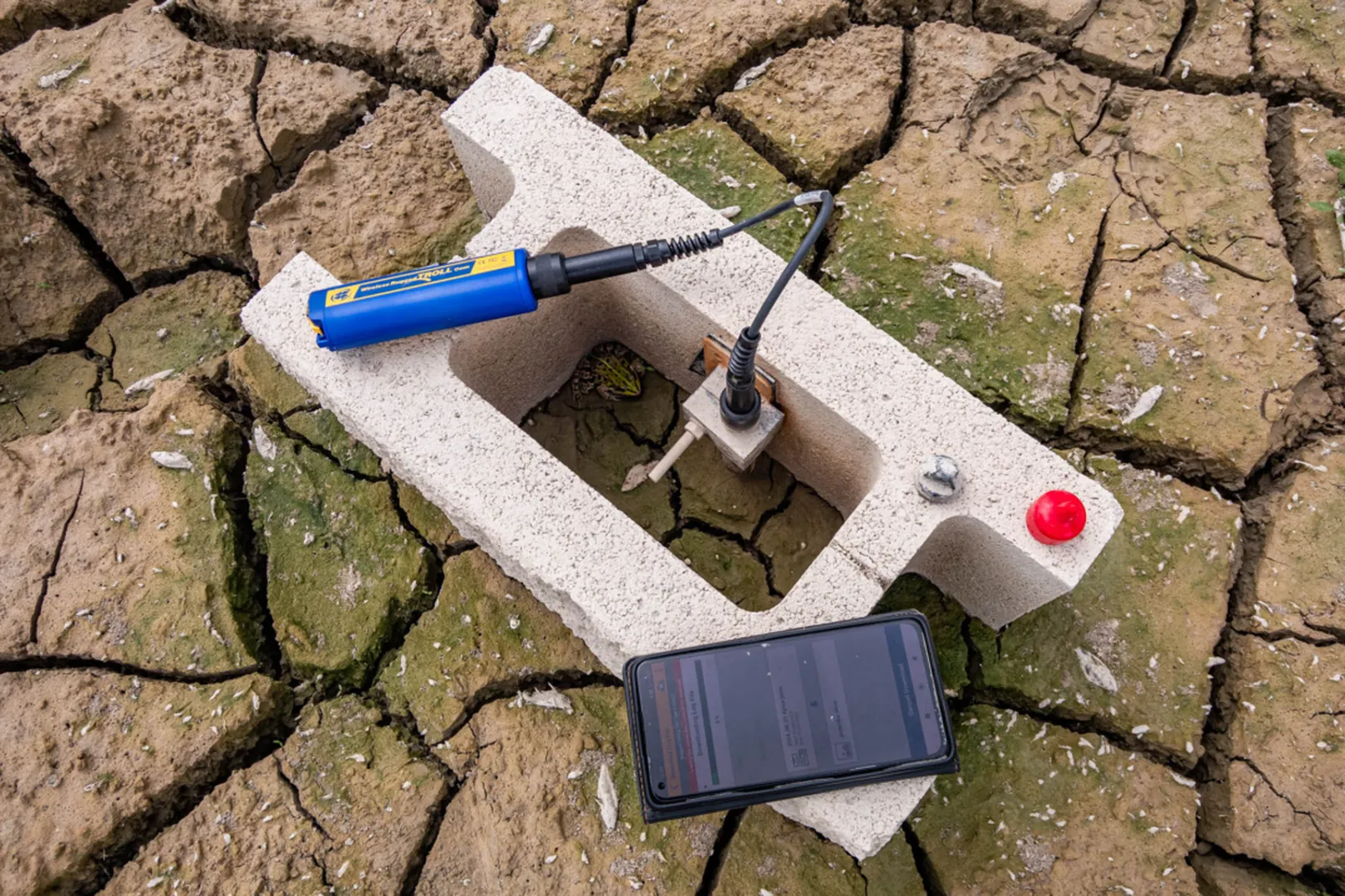 Merilna naprava za spremljanje vodostaja in temperature vode na suhem, razpokanem kraškem terenu: betonski blok z vgrajenim kovinskim senzorjem, priključenim na modro elektronsko napravo s kablom, poleg pametnega telefona, ki prikazuje podatke meritev.