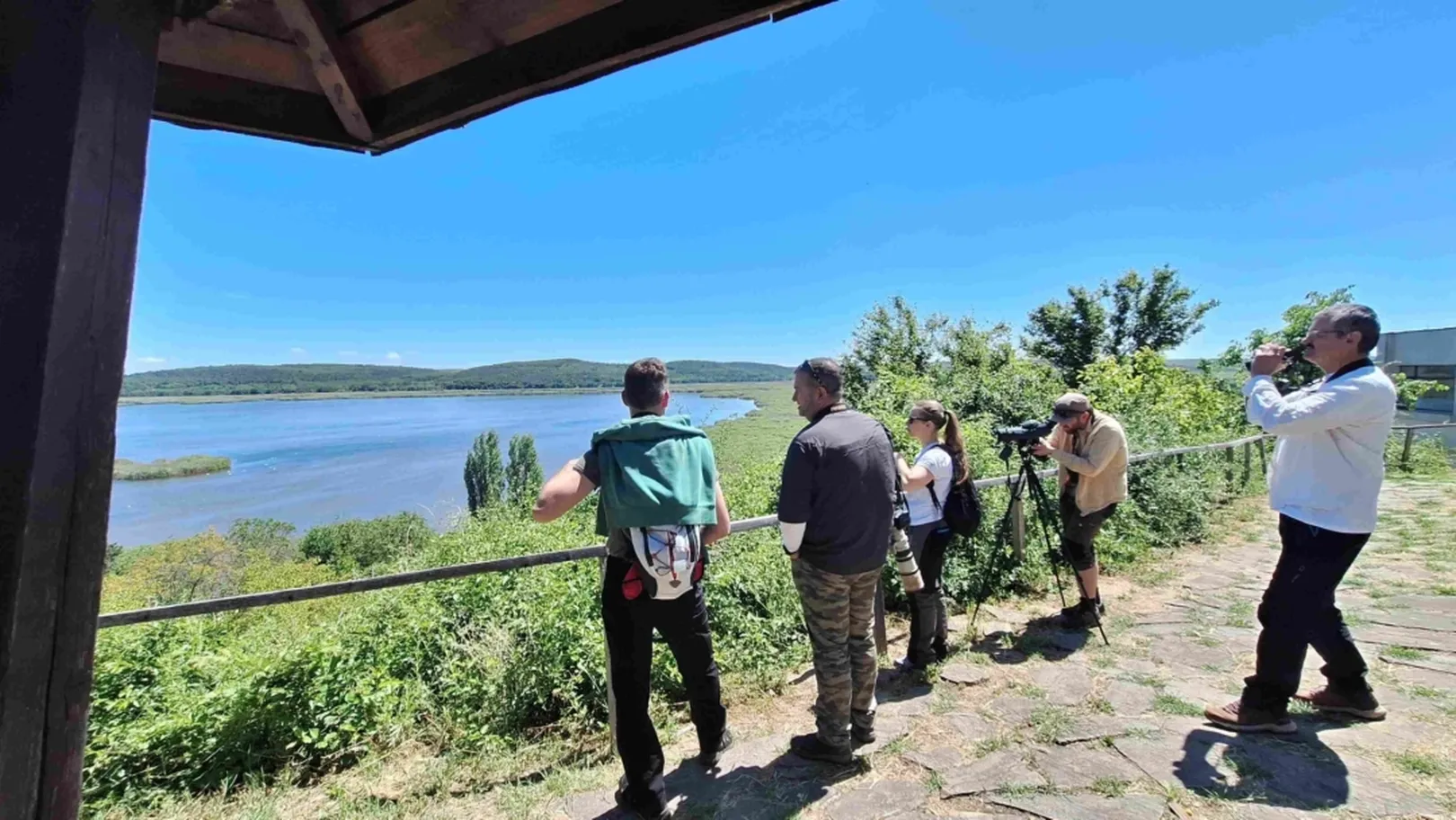 Skupina petih ljudi stoji na tlakovanem prostoru ob vodi, opazujejo naravo in pokrajino z daljnogledom, v ozadju zelenje in hribi pod jasnim modrim nebom