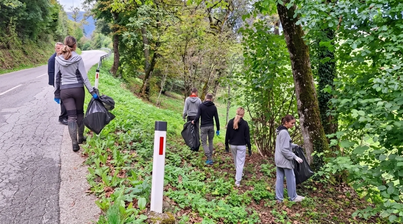 Skupina udeležencev ob cesti pobira odpadke na gozdnatem pobočju. Več oseb z zaščitnimi rokavicami nosi velike črne vreče za smeti, medtem ko se premikajo med drevesi in grmovjem ob robu ceste. V ozadju so vidni ovinek ceste, varnostni količki in gosto zelena vegetacija.