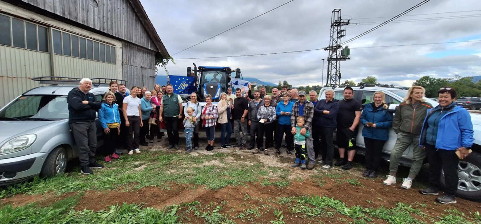 Skupinska fotografija udeležencev dogodka na dvorišču pred gospodarskim poslopjem. Zbrana skupina ljudi stoji med parkiranimi vozili in traktorjem z zastavo projekta LIFE. V ozadju so vidni električni drogovi, drevesa in oblačno nebo.