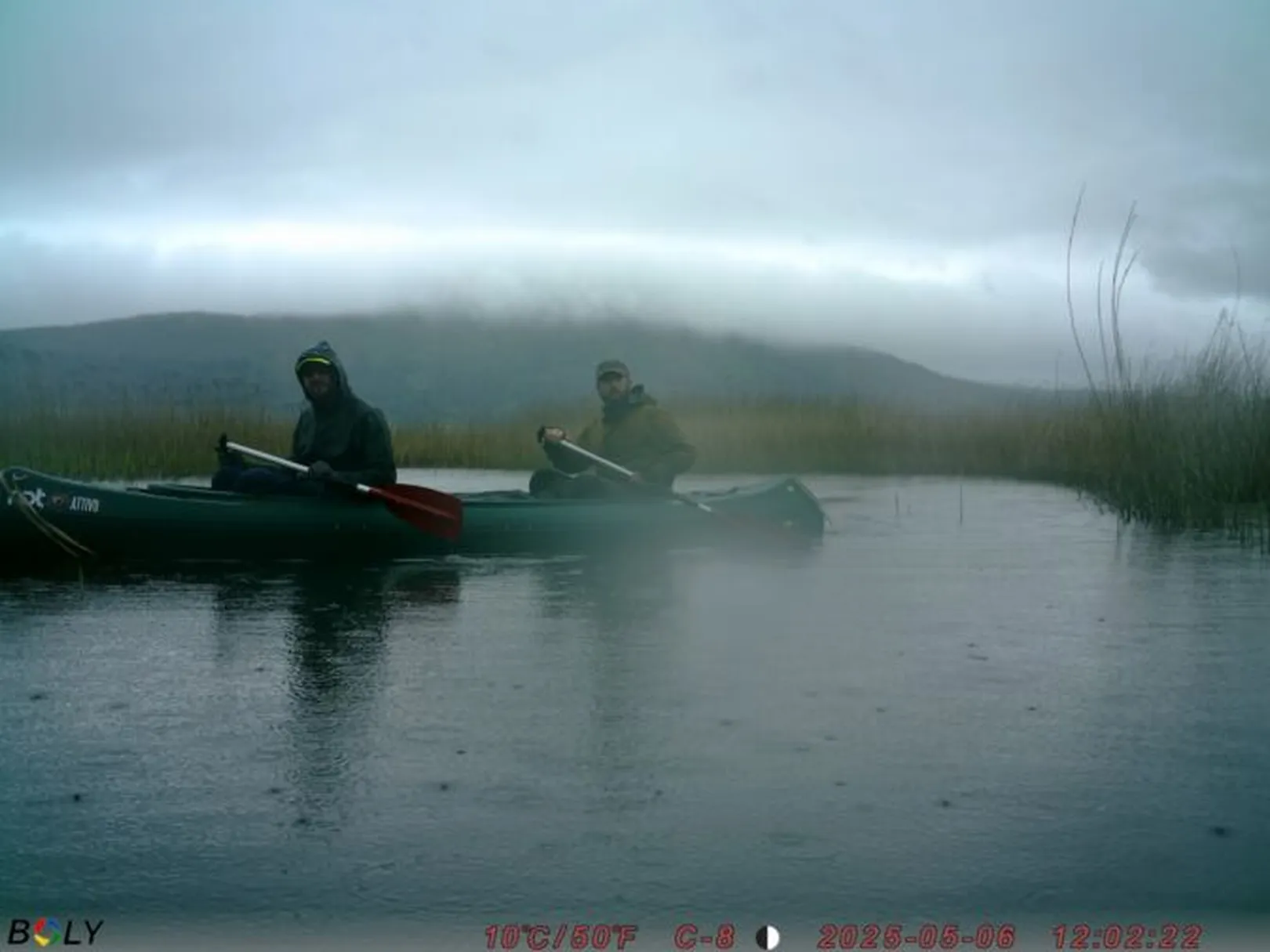 Dve osebi v kanuju na vodi, v ozadju trstičje in hribi pod meglenim, oblačnim nebom; prikazani so podatki o temperaturi in času.