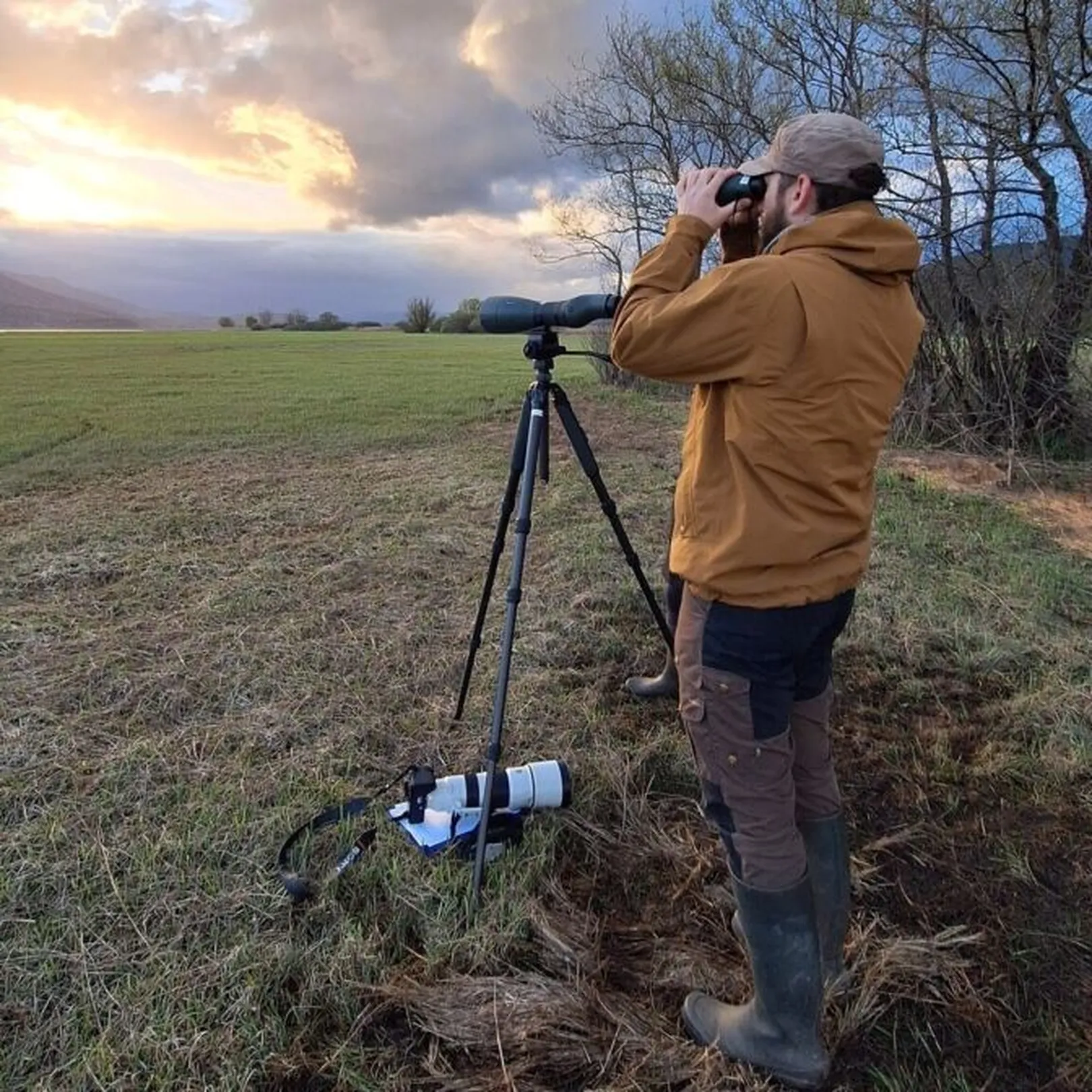 Oseba v rjavi jakni in kapi stoji na prostem ter opazuje z daljnogledom poleg stojala s kamero ali teleskopom; v ozadju polje, drevesa in delno oblačno nebo s sončnimi žarki.